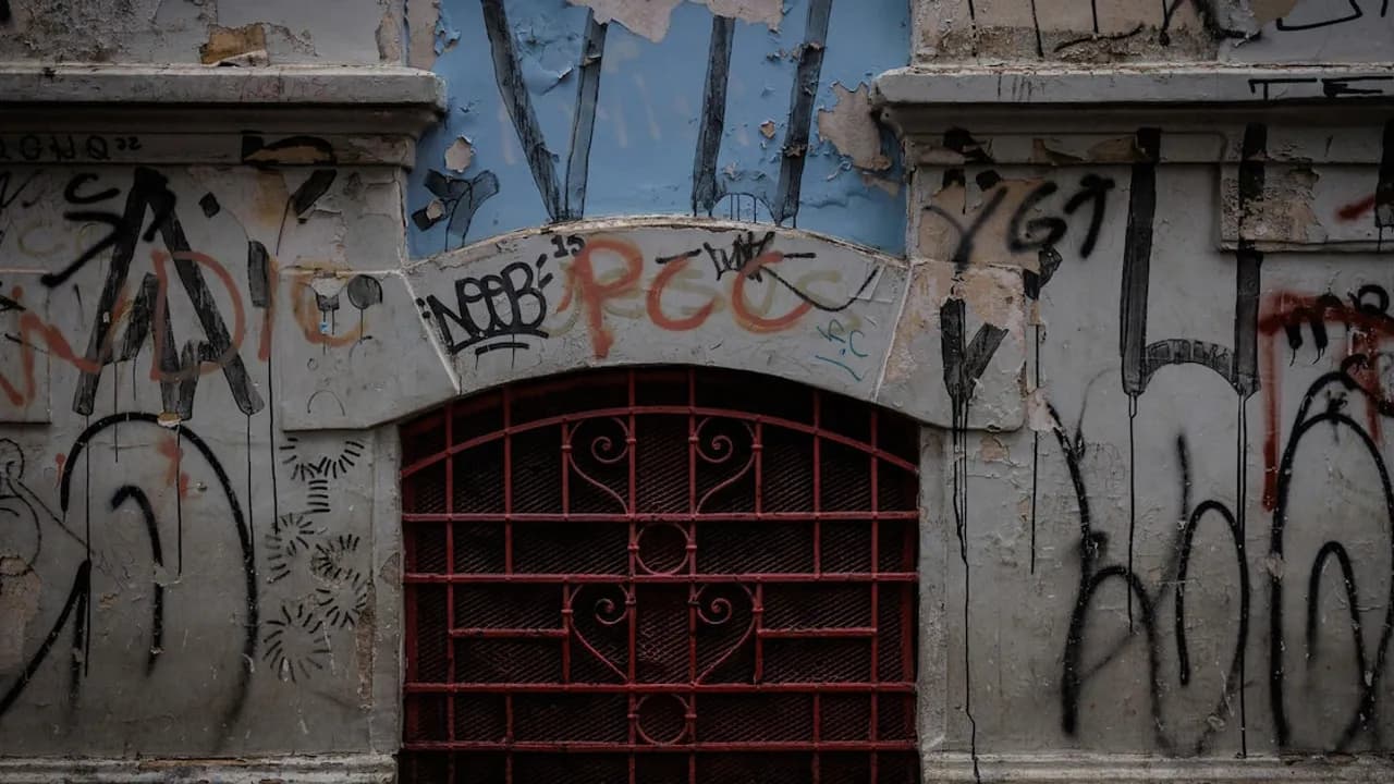 Pared antigua descascarada con grafitis y letras alusivas al PCC sobre un enrejado metálico curvo.