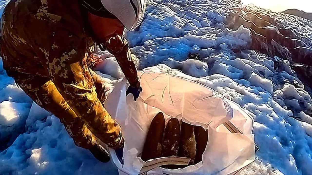 Militar con uniforme de camuflaje y casco revisa proyectiles de artillería dentro de una bolsa blanca sobre la nieve.