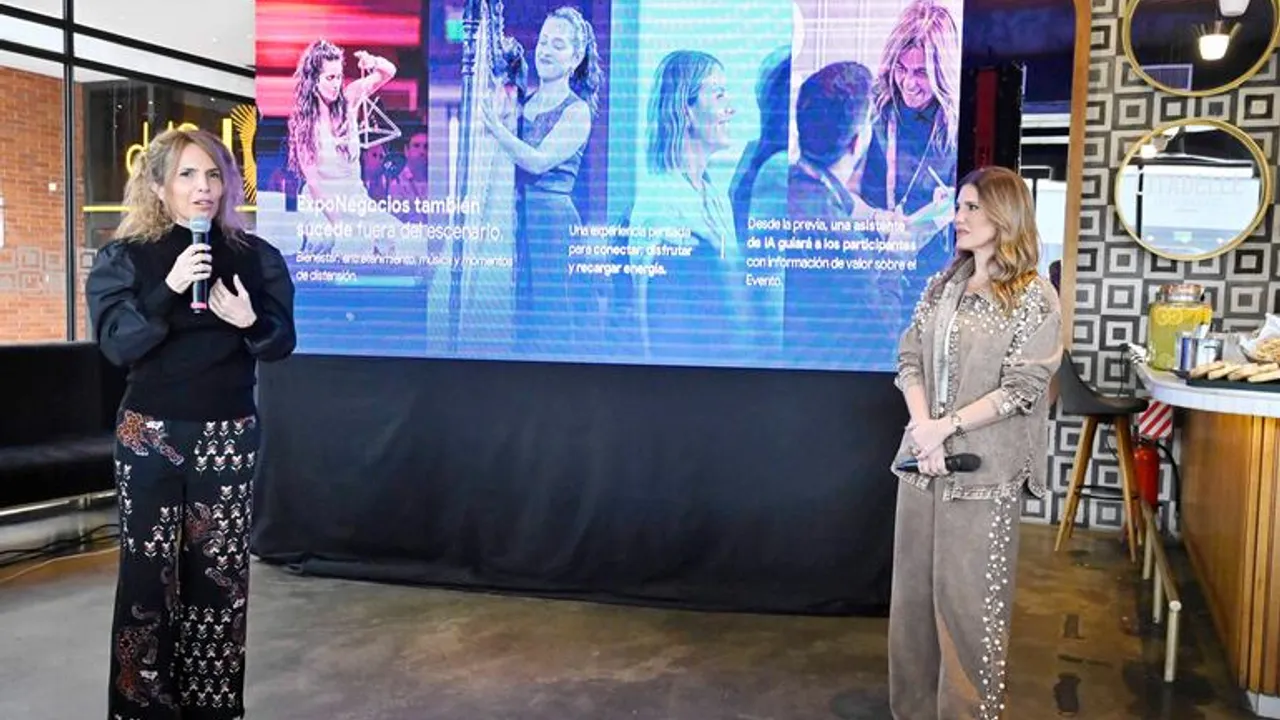 Dos mujeres con micrófonos presentan el evento frente a una pantalla digital con imágenes corporativas.