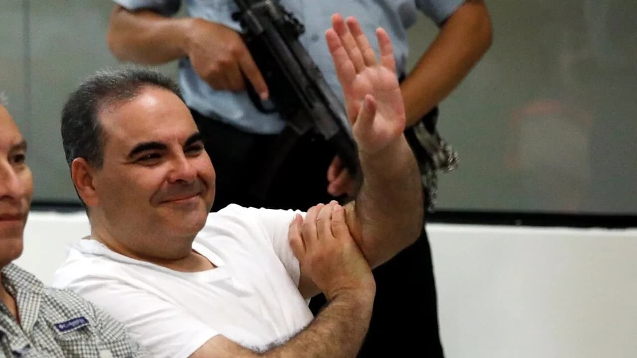 Elías Antonio Saca levanta una mano en un recinto custodiado por agentes de seguridad durante una comparecencia.