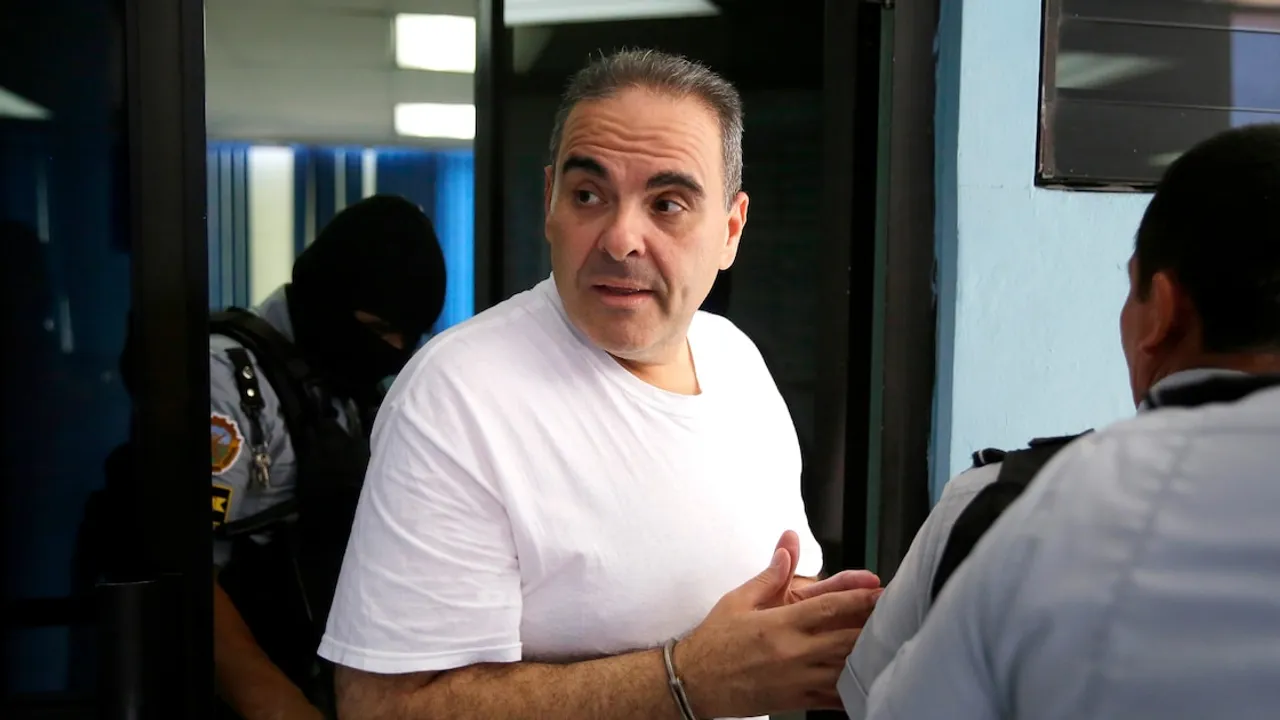 Hombre con camiseta blanca y esposas en las muñecas dialoga mientras es escoltado por agentes policiales.