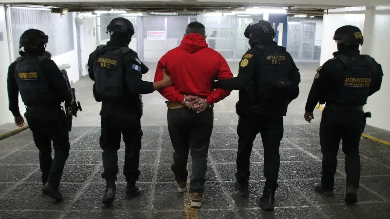 Agentes antinarcóticos con uniformes oscuros escoltan a un hombre esposado con campera roja por un pasillo interior.