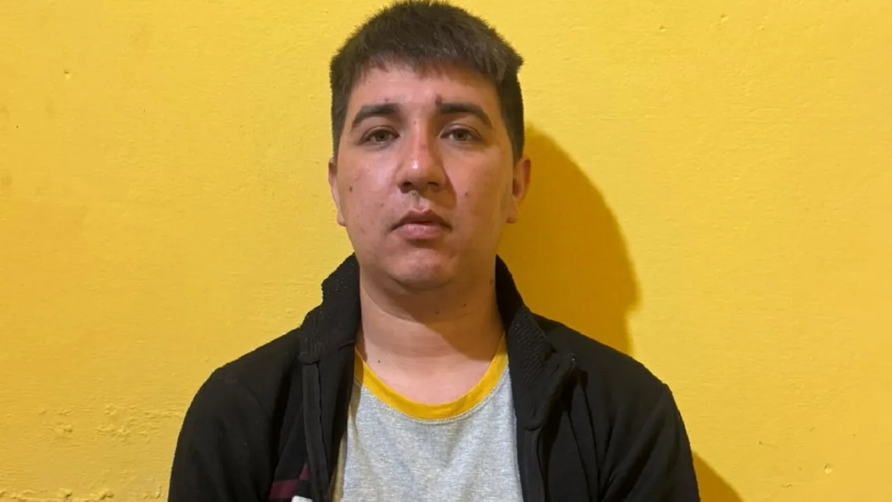 Hombre joven con campera oscura y remera gris posando de frente con la mirada seria ante una pared amarilla.