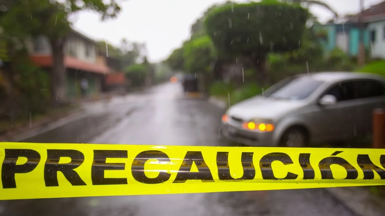 Cinta policial amarilla con la palabra precaución bloquea una calle asfaltada mojada por la lluvia con un auto de fondo.
