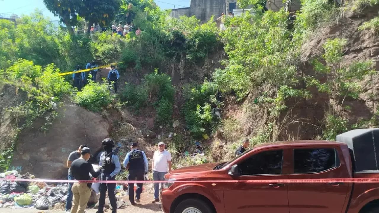 Agentes policiales y peritos inspeccionan un terreno escarpado con vegetación junto a una camioneta roja acordonada.