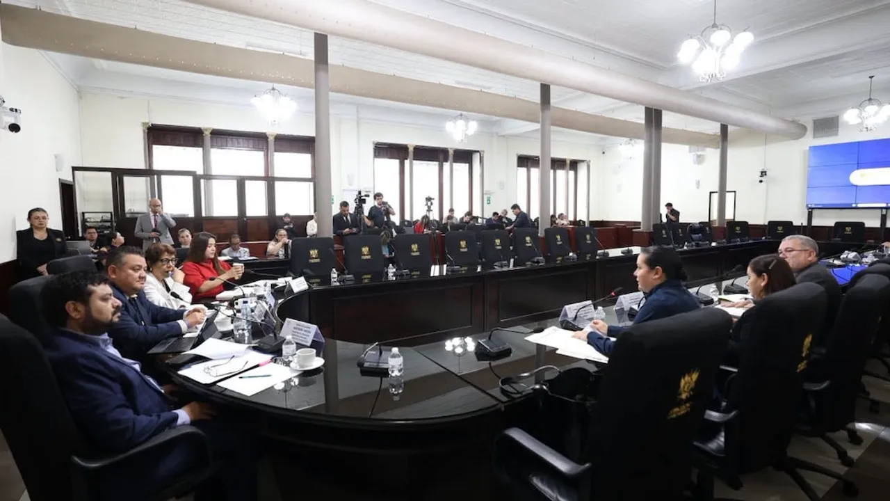 Funcionarios gubernamentales sentados alrededor de una mesa ovalada en una sala del Congreso con documentos y micrófonos.