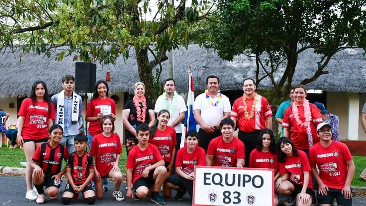 Grupo de jóvenes y docentes con remeras rojas y collares de flores posan frente a un cartel que dice Equipo 83.