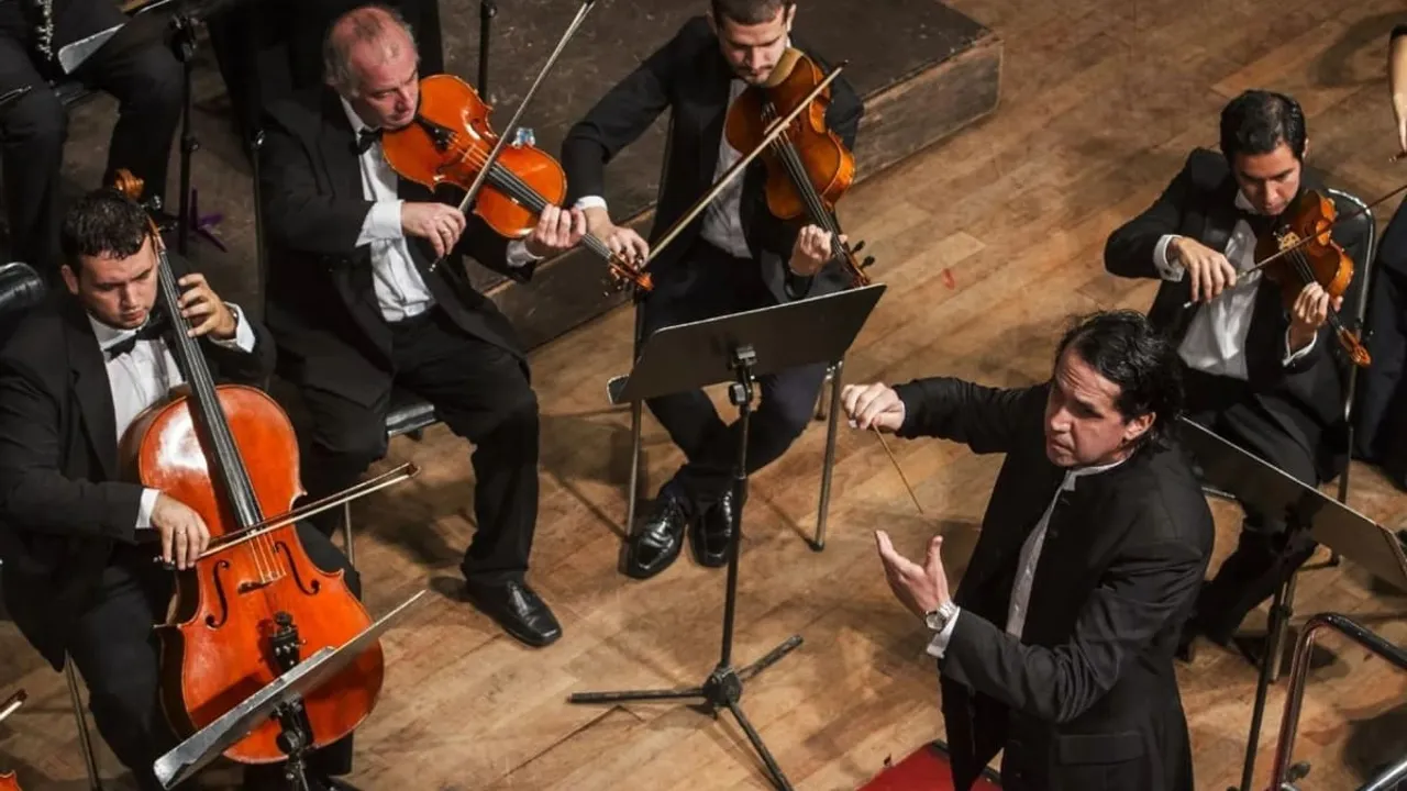 Músicos de orquesta tocan violines y chelo vestidos de gala bajo la conducción de un director que sostiene una batuta.
