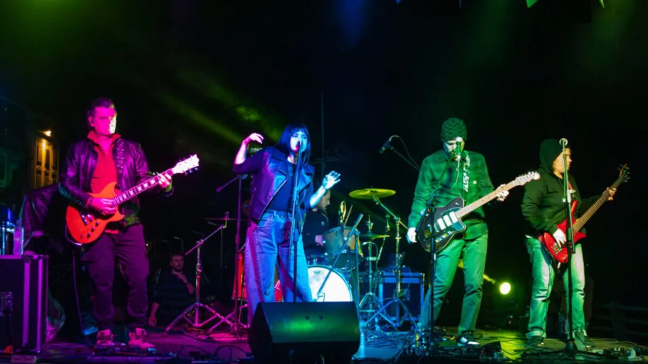Músicos de una banda tocan guitarras y cantan en un escenario con reflectores de luces de color verde y violeta.