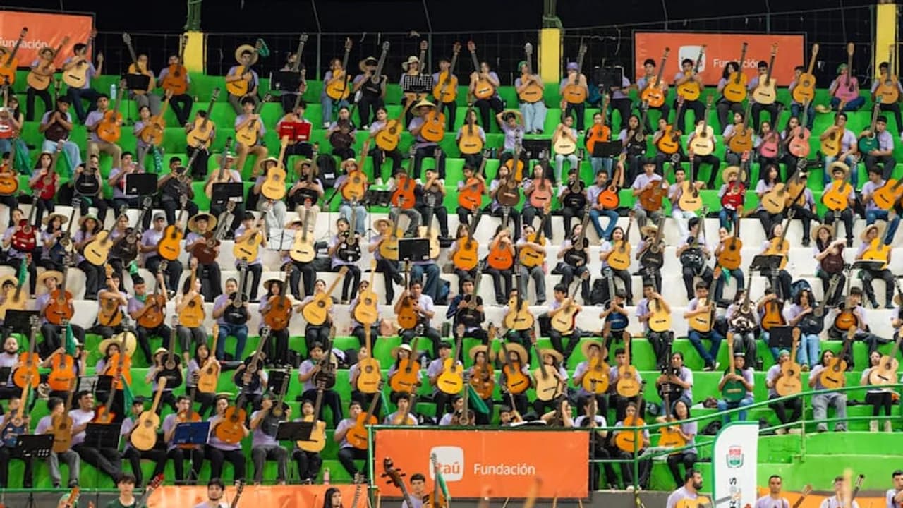 Decenas de jóvenes músicos sentados en gradas sostienen guitarras acústicas durante un concierto orquestal