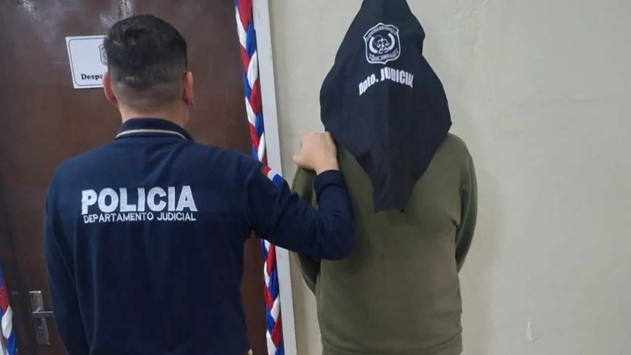 Un agente policial custodia a un hombre encapuchado que viste buzo verde y permanece de espaldas contra una pared.