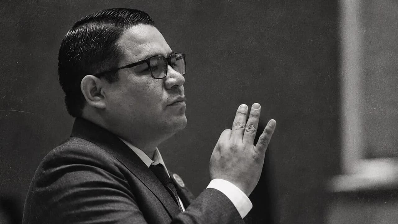 Hombre de traje y anteojos de perfil en blanco y negro levantando tres dedos con la mano derecha en un recinto oficial.