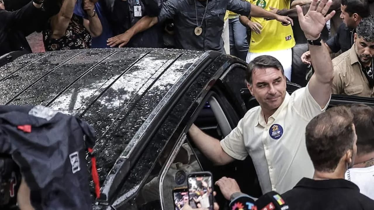 Flávio Bolsonaro saluda con la mano levantada desde un automóvil negro ante una multitud de personas.
