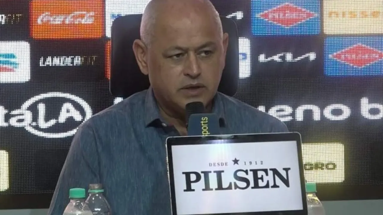 Hombre de cabello canoso y camisa azul habla frente a un micrófono en una sala de prensa con pantallas publicitarias