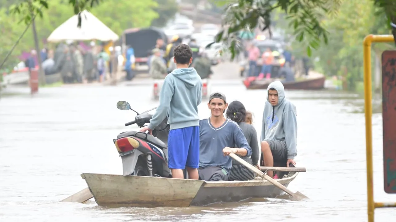 Personas a bordo de un bote de madera navegan por una calle inundada con una motocicleta encima.