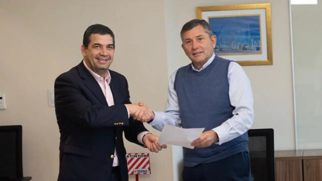 Dos hombres con vestimenta formal estrechan sus manos mientras sostienen un documento impreso dentro de una oficina.