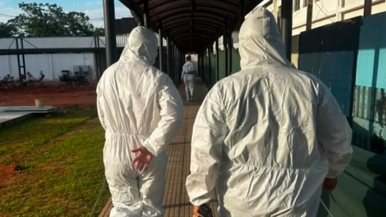 Dos personas con trajes blancos de protección química caminan por un pasillo techado en una planta industrial.