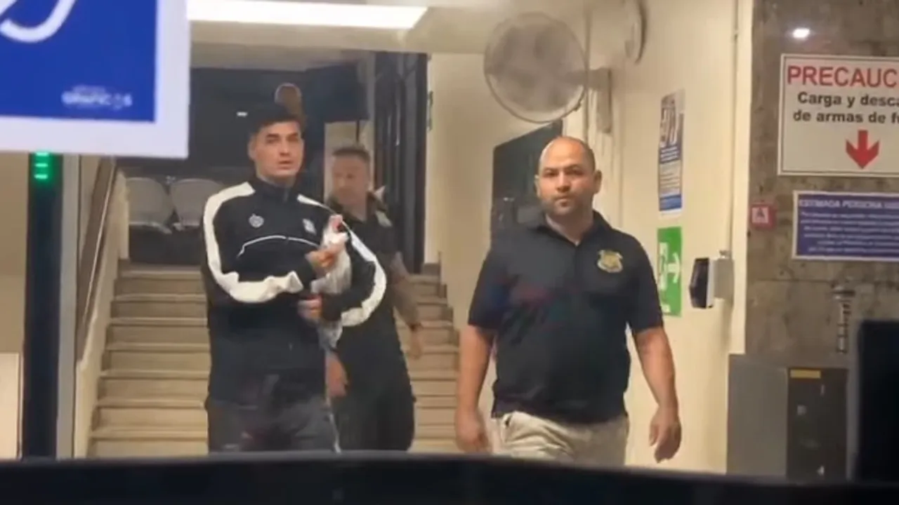 Un hombre con indumentaria deportiva negra y blanca baja por una escalera escoltado por personal de seguridad.