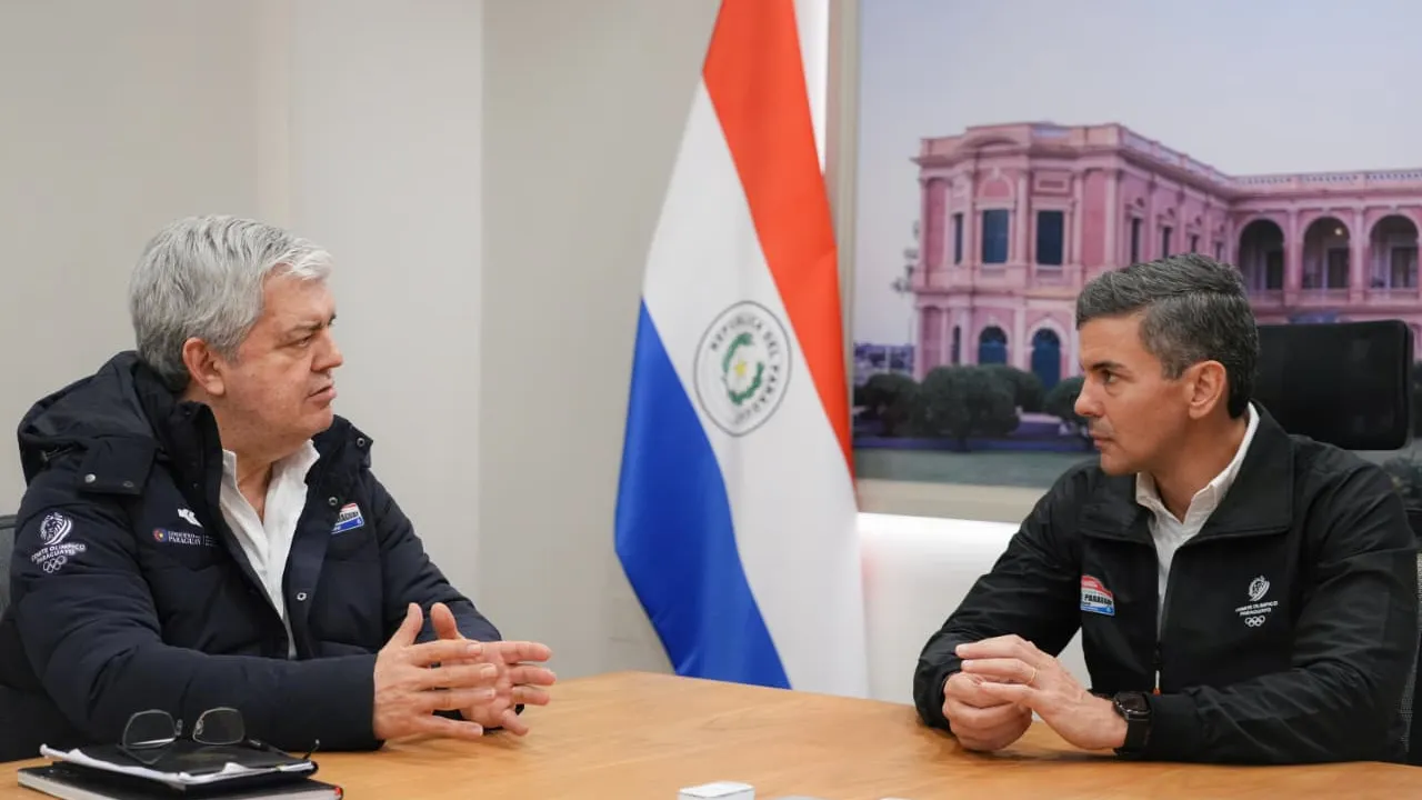 Enrique Riera y Santiago Peña conversan sentados frente a una mesa con la bandera paraguaya de fondo