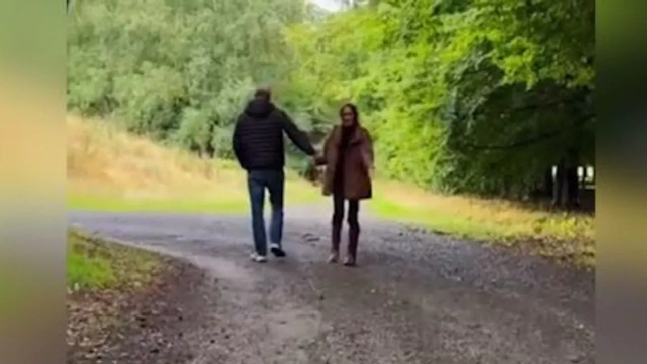 Un hombre y una mujer caminan de espaldas tomados de la mano por un sendero arbolado de tierra.