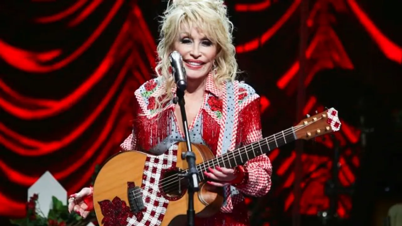Dolly Parton sonríe en el escenario sosteniendo una guitarra acústica con detalles rojos y un micrófono frente a ella.