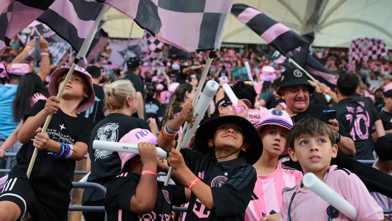Niños y aficionados con camisetas rosadas agitan banderas y bastones en las gradas de un estadio deportivo.