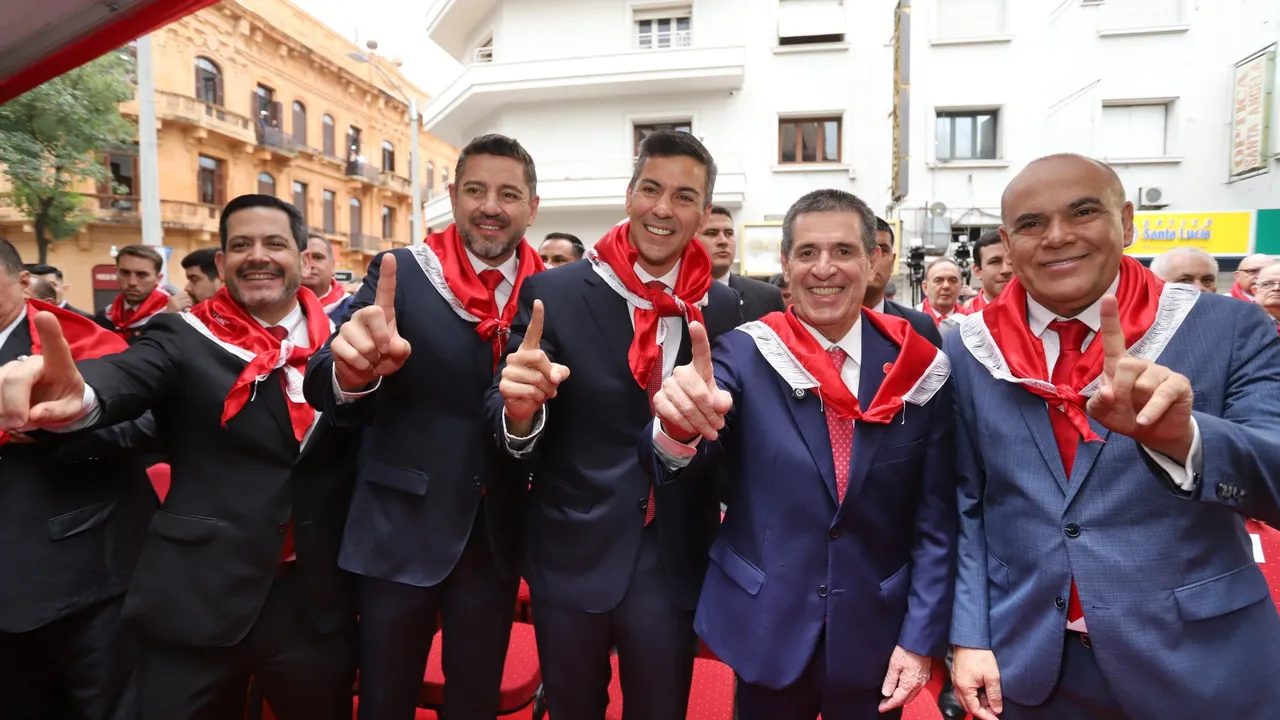 Autoridades y dirigentes con pañuelos rojos al cuello posan sonrientes y con el dedo índice en alto en un acto público.