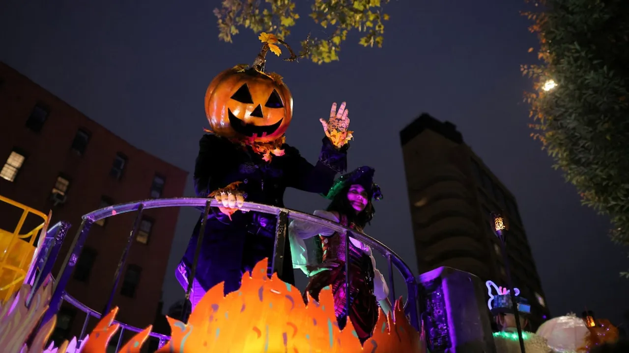 Persona disfrazada con una gran cabeza de calabaza saluda desde una carroza festiva en un desfile nocturno.