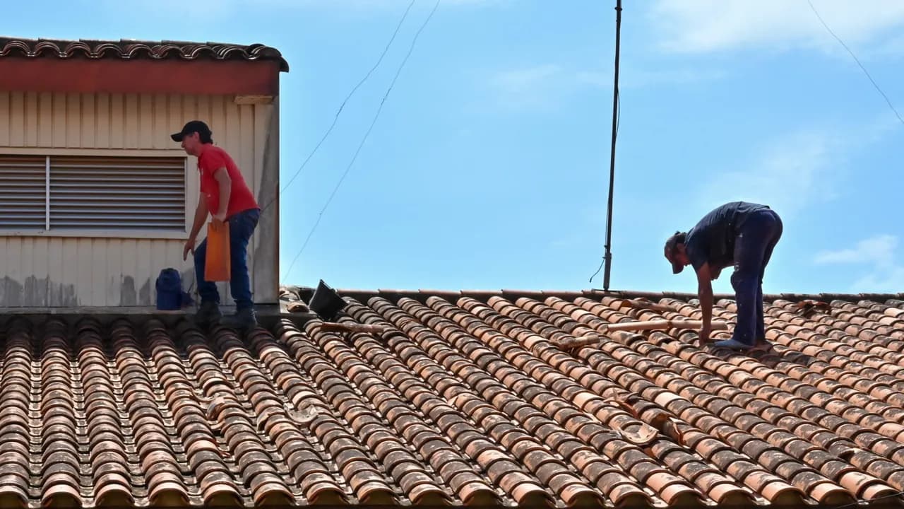 Dos operarios con gorra reparan las tejas coloniales dañadas sobre el tejado de un pabellón bajo el cielo despejado.