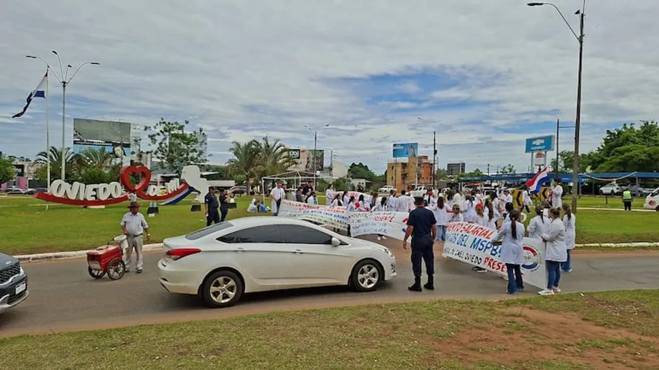 Manifestantes con batas blancas sostienen pancartas y bloquean el tránsito frente a una rotonda urbana con vehículos.