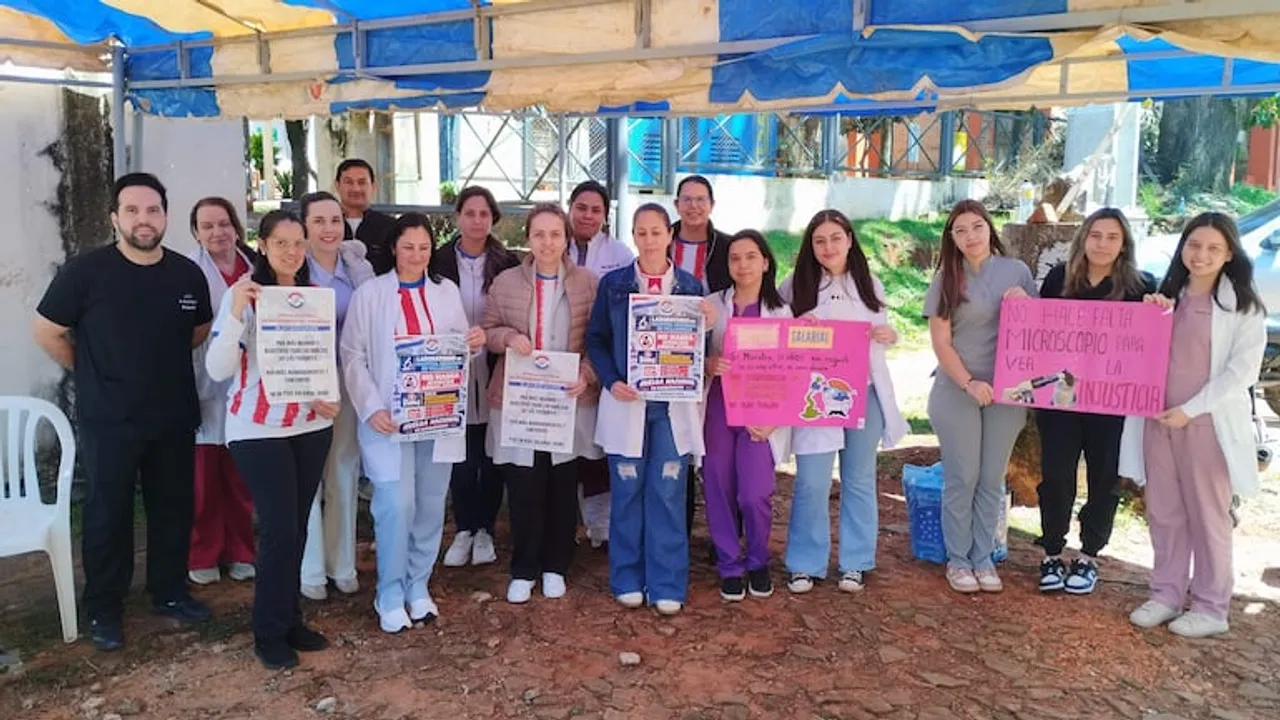 Grupo de bioquímicos y personal de blanco bajo una carpa sosteniendo carteles de protesta en Guairá.
