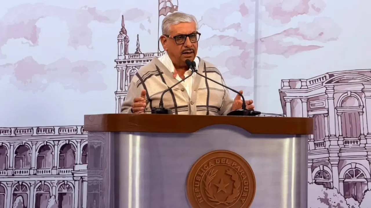 Isaías Fretes con anteojos y abrigo habla detrás de un podio con el escudo de la República del Paraguay.
