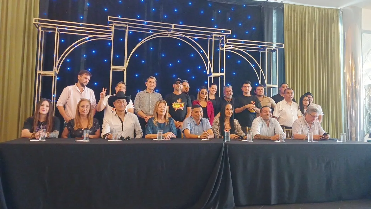Grupo de personas en una conferencia de prensa sentadas y de pie frente a una mesa larga con fondo oscuro decorado.