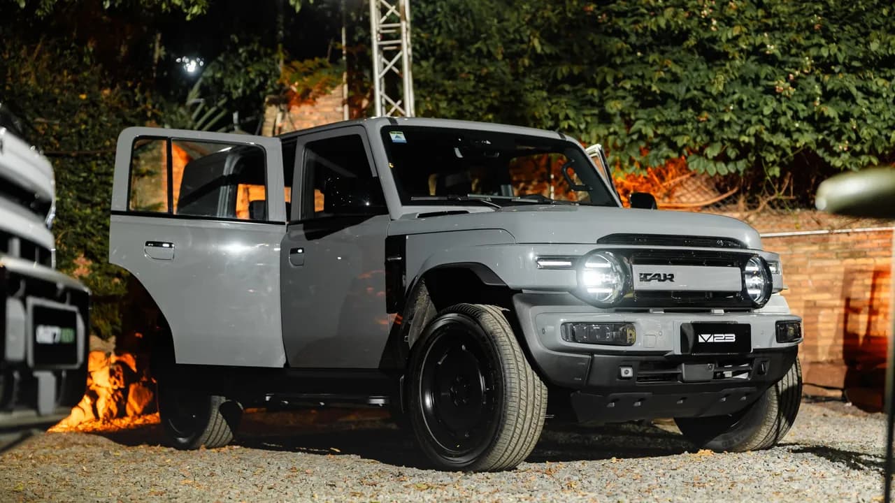 Camioneta todoterreno gris con luces delanteras redondas encendidas y una puerta abierta sobre terreno de gravilla