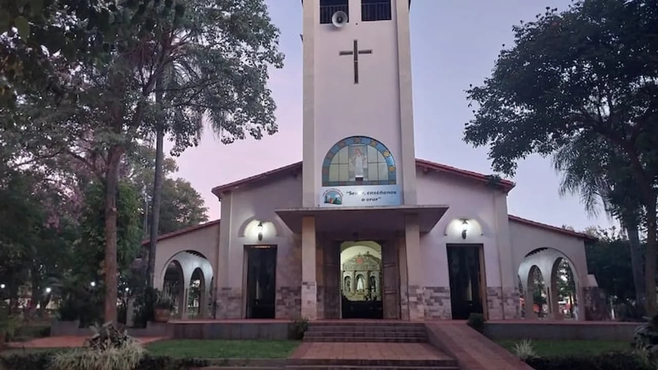 Fachada de la iglesia con torre campanario central, cruz superior y arcos laterales rodeada de árboles al atardecer.