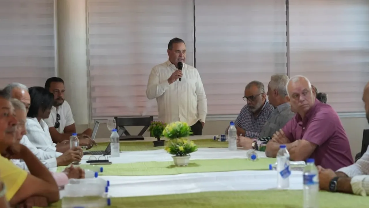 Hombre de pie con camisa blanca habla por micrófono frente a varias personas sentadas en una mesa de reunión.