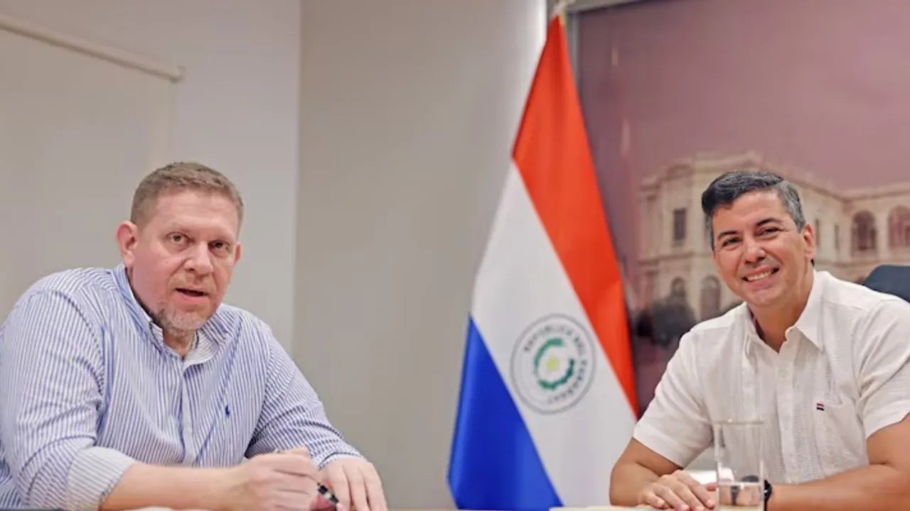 Eddie Jara y Santiago Peña conversan sentados frente a una mesa junto a una bandera paraguaya.