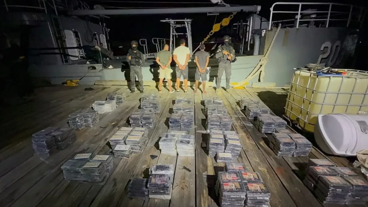 Agentes armados custodian a tres hombres detenidos junto a decenas de paquetes de droga decomisada en una cubierta.