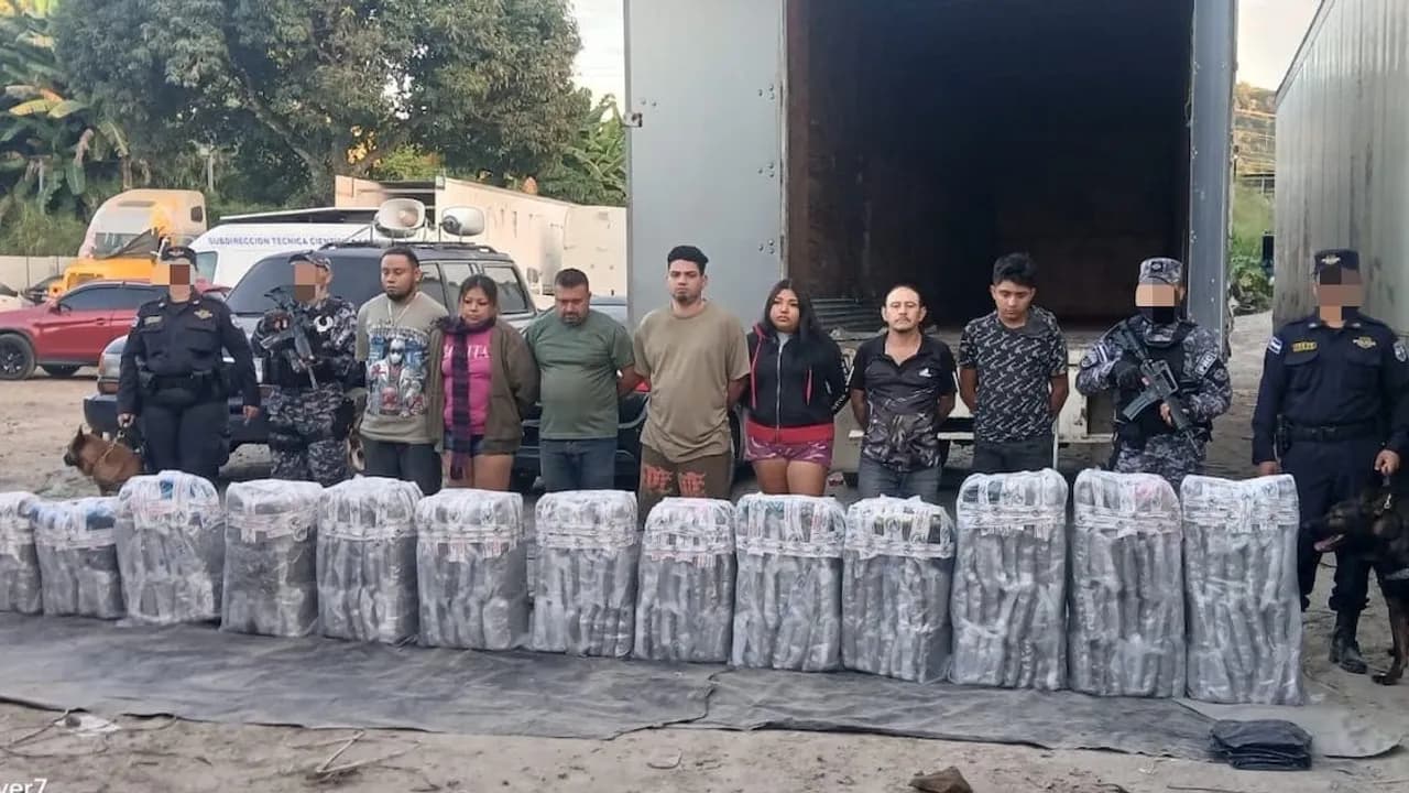 Siete personas detenidas y agentes armados posan frente a un camión detrás de paquetes asegurados en el suelo.