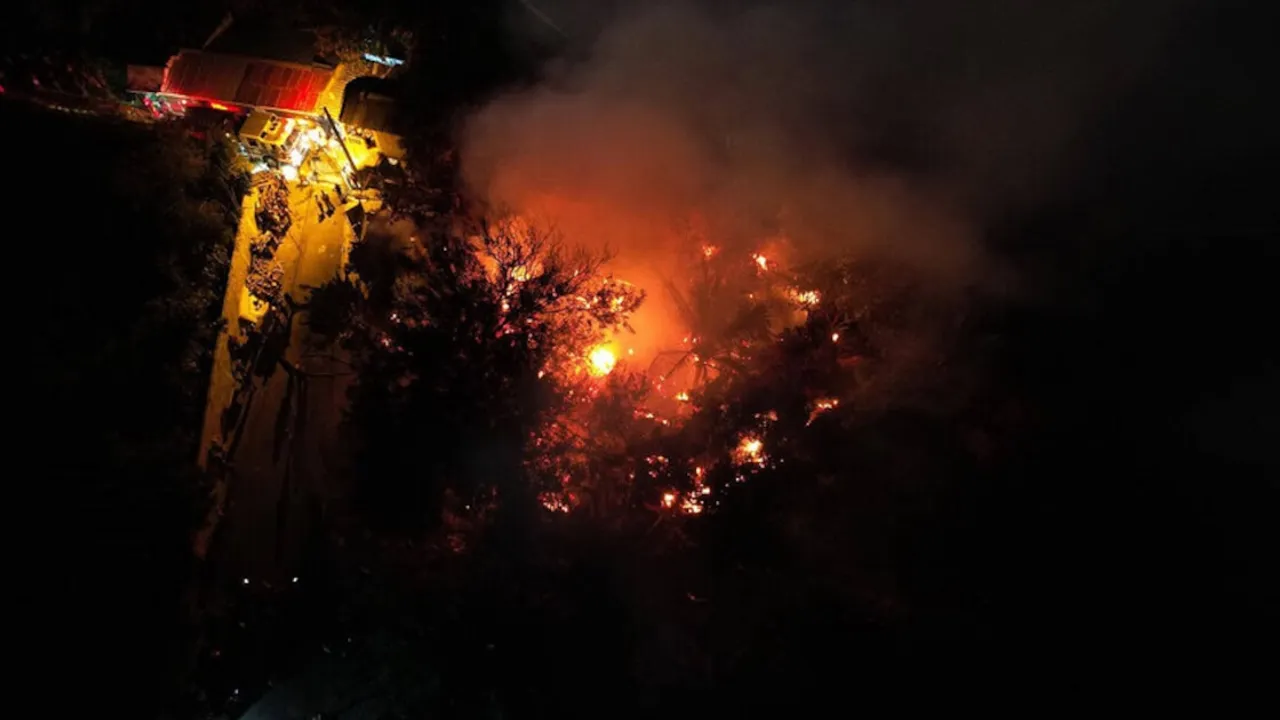 Vista aérea nocturna de un incendio forestal y estructural con llamas intensas y humo en medio de árboles.
