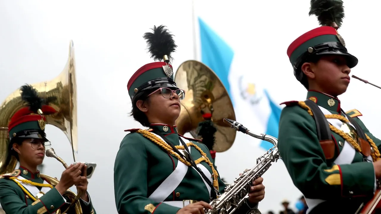 Integrantes de una banda escolar tocan saxofón y otros instrumentos de viento con uniformes de gala y banderas detrás.