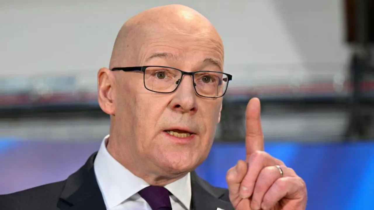 John Swinney, con lentes y traje oscuro, gesticula con el dedo índice levantado frente a un micrófono.