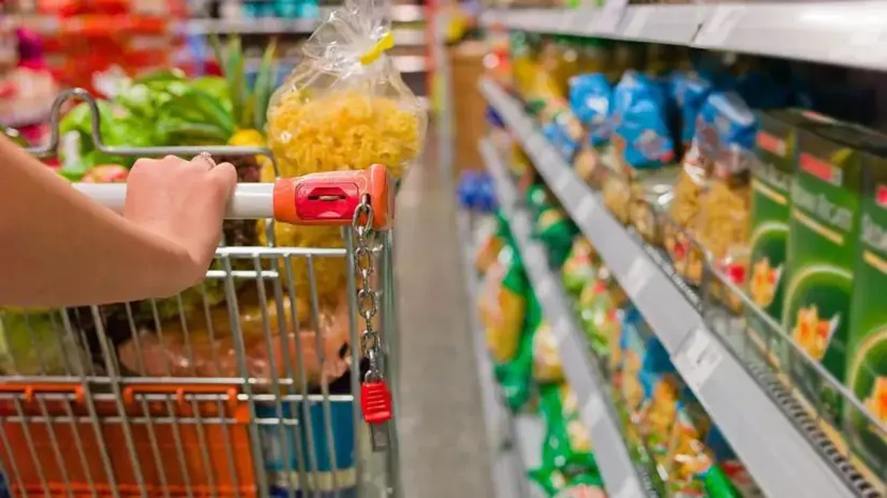 Mano de una persona empujando un carrito de supermercado por un pasillo con góndolas repletas de alimentos.