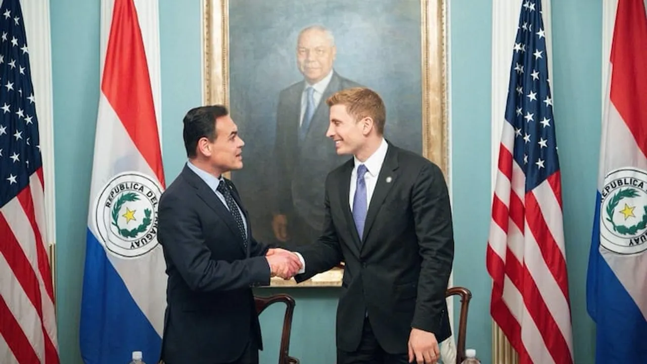 Dos autoridades estrechan la mano en un despacho oficial frente a banderas de Paraguay y Estados Unidos.