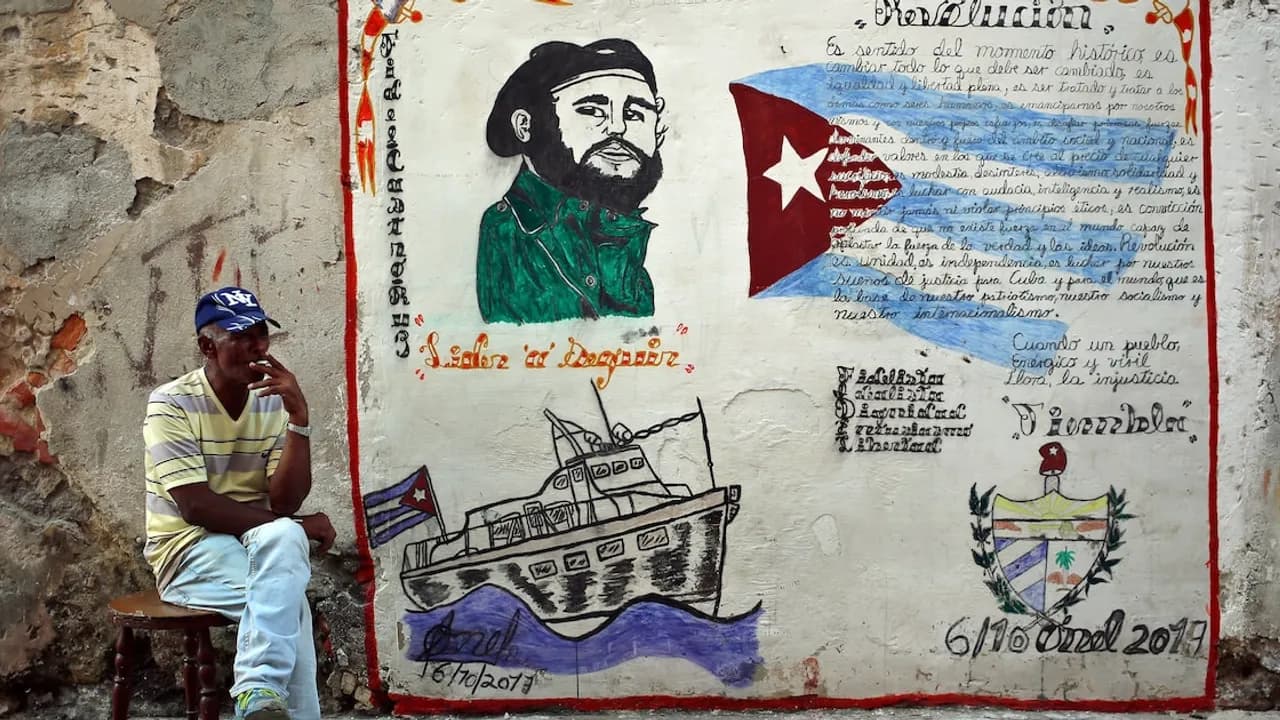 Hombre sentado frente a un mural desgastado con el rostro de Fidel Castro, el barco Granma y la bandera cubana.