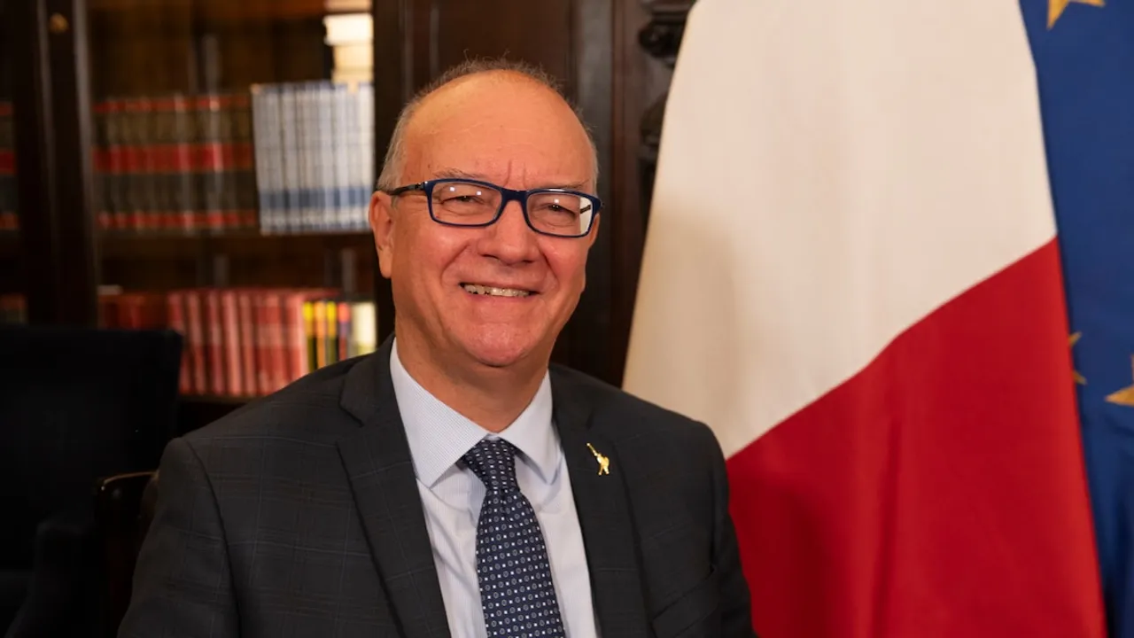 Giuseppe Valditara con traje oscuro, corbata estampada y anteojos, sonriendo ante banderas institucionales.