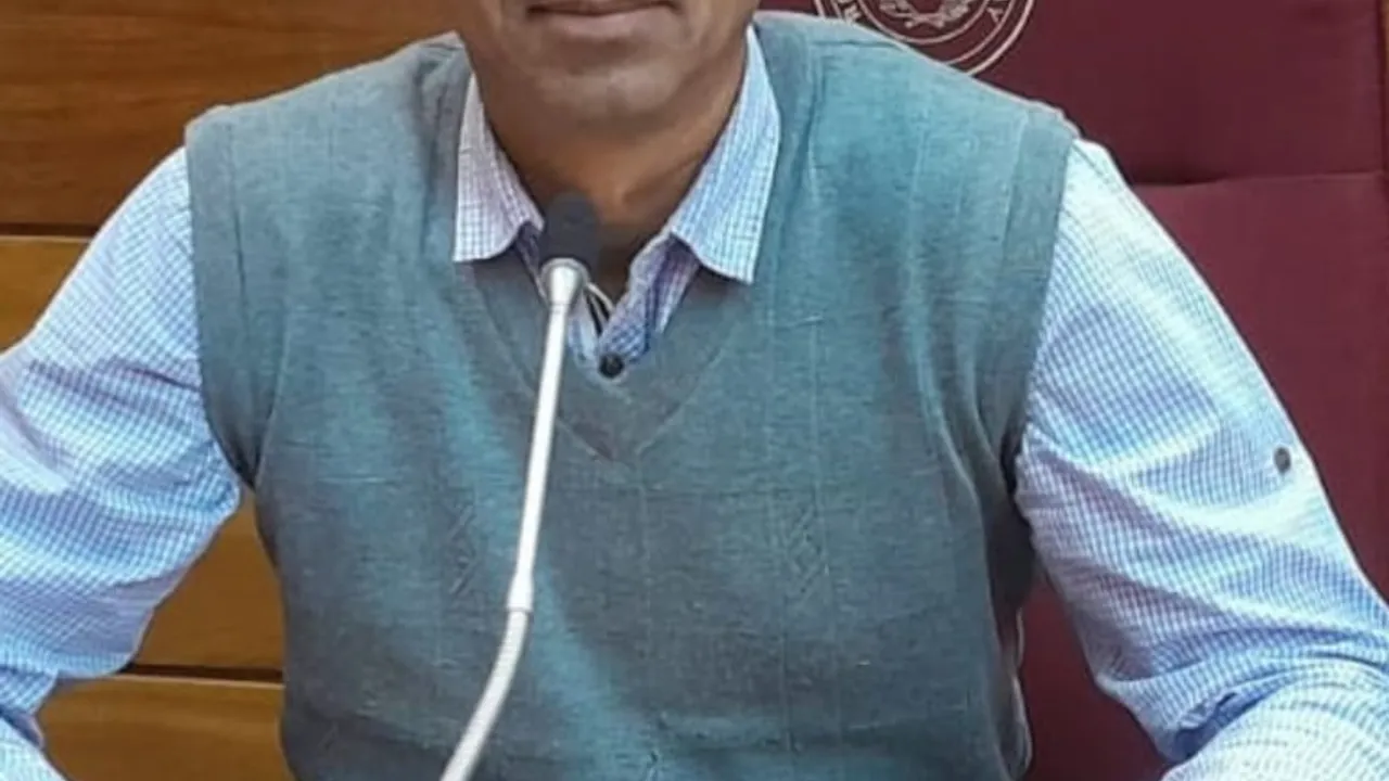 Hombre con chaleco gris y camisa a cuadros sentado frente a un micrófono en una mesa institucional de madera.