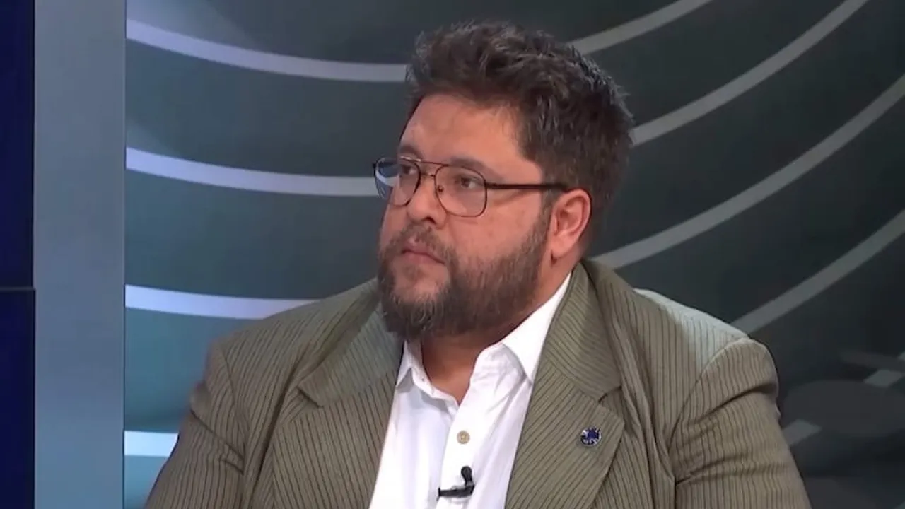 Hombre con barba y lentes, vestido con saco a rayas y camisa blanca, durante una entrevista en un set televisivo.