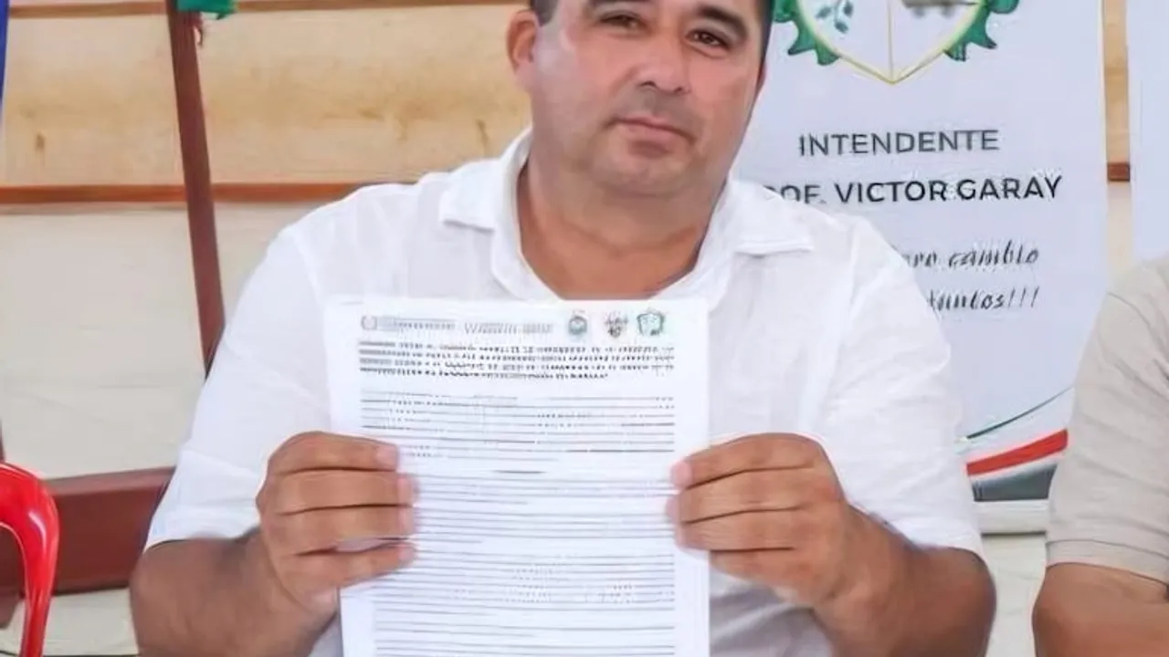 Hombre de camisa blanca sentado sosteniendo una hoja impresa con texto frente a una pancarta institucional.