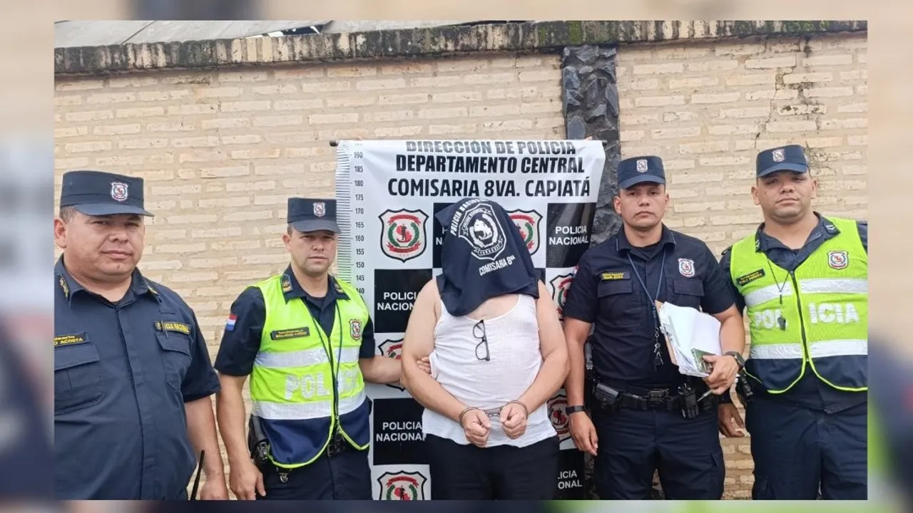 Agentes policiales custodian a un hombre esposado y con el rostro cubierto frente a un cartel de la comisaría de Capiatá.